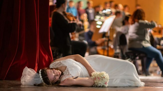 Im Vordergrund liegt die Darstellerin der Juliette in einem weißen Brautkleid im Sterben. Im Hintergrund sieht man verschwommen die Musiker und die Chöre.