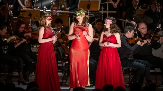 Im Vordergrund sind die Darstellerinnen der Juliette, Amelie und Antonia zu sehen. Im Hintergrund sieht man die Musiker des Loh-Orchesters.